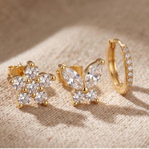 May include: A collection of three gold-coloured earrings. One pair is a flower stud with clear gemstones, another is a butterfly stud with clear gemstones, and the third is a small hoop earring with clear gemstones.
