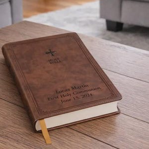 May include: A brown leather-bound Bible with gold-colored page edges and a matching bookmark. The cover features a cross and the words "Holy Bible" above the name "Lucas Martin" and the date "June 15, 2024". The book is on a wooden table.