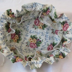 May include: A round, floral fabric bowl with a ruffled edge. The bowl features a cream-colored background with a pattern of pink roses, yellow flowers, and blue vines. The ruffled edge adds a decorative touch to the bowl.