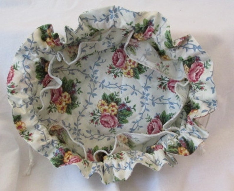May include: A round, floral fabric bowl with a ruffled edge. The bowl features a cream-colored background with a pattern of pink roses, yellow flowers, and blue vines. The ruffled edge adds a decorative touch to the bowl.