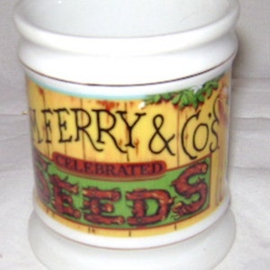 May include: A white ceramic jar with a cylindrical body and a flared rim. The jar features a label with the text "FERRY & CO'S CELEBRATED SEEDS" in black and red lettering against a yellow and green background, with a wood-grain design.