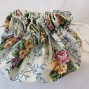 May include: A small, gathered drawstring pouch with a floral pattern. The fabric is cream-colored with a design of pink roses, yellow flowers, and blue vines. White satin ribbon forms the drawstring.
