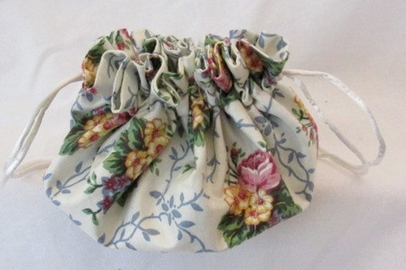 May include: A small, gathered drawstring pouch with a floral pattern. The fabric is cream-colored with a design of pink roses, yellow flowers, and blue vines. White satin ribbon forms the drawstring.