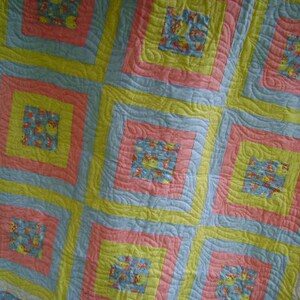 May include: A handmade quilt with a repeating square pattern. The quilt features squares of light blue, yellow, and pink, with a patterned center square. The quilt's design includes a repeating pattern of small, colorful figures.