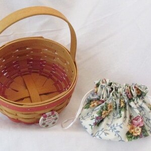 May include: A woven basket with a wooden handle and a floral drawstring pouch. The basket is light brown with a red interior and trim. The pouch is white with a floral pattern in pink, yellow, and blue.