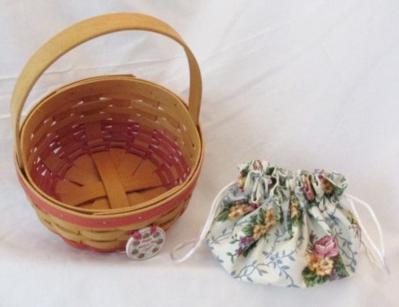 May include: A woven basket with a wooden handle and a floral drawstring pouch. The basket is light brown with a red interior and trim. The pouch is white with a floral pattern in pink, yellow, and blue.