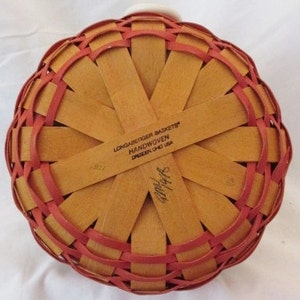 May include: A handwoven basket with a round base, featuring a woven pattern of light brown and red strips. The text "LONGABERGER BASKETS HANDWOVEN DRESDEN OHIO USA" is visible. The basket has a white handle.