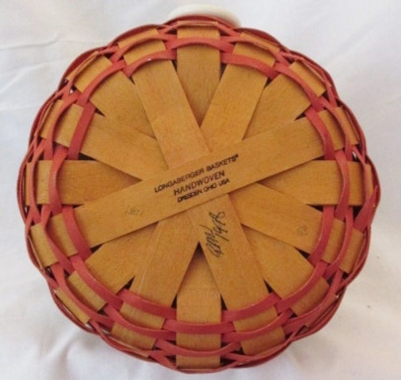 May include: A handwoven basket with a round base, featuring a woven pattern of light brown and red strips. The text "LONGABERGER BASKETS HANDWOVEN DRESDEN OHIO USA" is visible. The basket has a white handle.