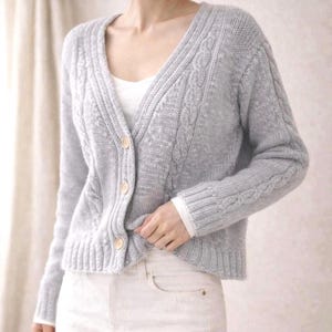 Può includere: Un cardigan grigio chiaro a maglia a trecce con scollo a V e bottoni in legno. Il maglione presenta un motivo a trecce strutturato sulle maniche e sui pannelli anteriori. Il cardigan è abbinato a un top bianco e jeans bianchi.