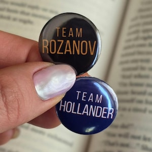 May include: Two round buttons with text. One button is black with "TEAM ROZANOV" in gold lettering. The other button is dark blue with "TEAM HOLLANDER" in white lettering. The buttons are held in front of an open book.