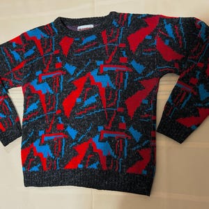 Pode incluir: Um suéter de manga comprida com gola redonda em cinza carvão escuro com um padrão geométrico repetitivo em vermelho vibrante e azul brilhante. A peça de malha tem gola, punhos e bainha canelados com um design angular e abstrato.