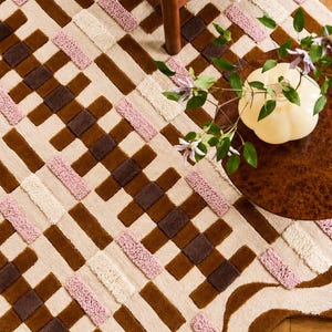 Op de afbeelding: Een patroonkleed in bruin, beige en roze tinten. Het kleed heeft een geweven ontwerp met rechthoekige blokken en een gebogen rand. Een houten stoel en een tafel met een vaas bloemen zijn gedeeltelijk zichtbaar.