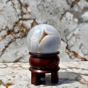 Peut inclure: Une sphère en agate bleu clair et blanche avec un intérieur brun, présentée sur un support en bois rouge foncé. L'agate présente un motif craquelé. Le support a une base ronde et une base carrée en forme de piédestal.