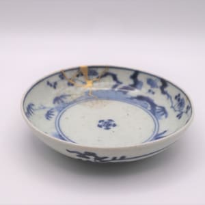 Op de afbeelding: Een ronde, gebroken witte keramische schaal met blauwe bloempatronen en een blauwe rand. De schaal is gerepareerd met goudkleurige kintsugi, een Japanse kunstvorm. Op een witte achtergrond.