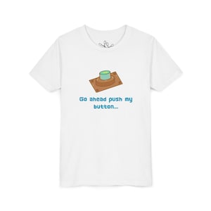 May include: A white short-sleeve t-shirt featuring a graphic of a brown rectangular base with a light blue circular button on top. Below the graphic, the text reads "Go ahead push my button..." in a blue, pixelated, blocky font style.