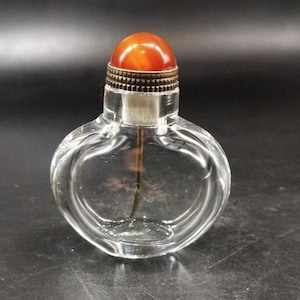 May include: A clear, flattened, oval-shaped glass snuff bottle with a rounded base. The bottle features a metallic, textured collar and a smooth, polished, dome-shaped orange stone stopper. A thin, dark applicator rod extends from the stopper.