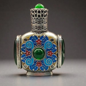May include: A decorative perfume bottle featuring a silver-toned metal body with intricate filigree patterns. The centre is adorned with a large green cabochon stone surrounded by blue enamel floral motifs with small red flower accents.