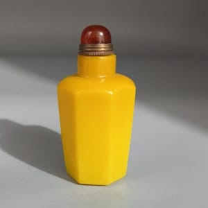May include: A bright yellow hexagonal snuff bottle with a smooth, opaque finish. The bottle features a narrow neck topped with a metallic, ribbed collar and a rounded, translucent dark reddish-brown stopper that sits securely on the top opening.