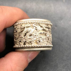 Könnte beinhalten: Ein zylindrischer Ring aus silberfarbenem Metall mit aufwendigen, durchbrochenen Reliefschnitzereien eines Drachen inmitten wirbelnder Wolken. Das Band ist an den oberen und unteren Rändern mit einer dekorativen Perlenverzierung versehen.