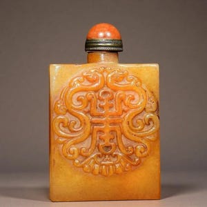 May include: A rectangular yellow jade snuff bottle featuring a central circular relief carving of a stylized dragon motif. The bottle is topped with a rounded orange stone stopper set in a dark metal band, creating a contrast with the smooth stone body.