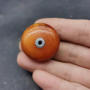 Op de afbeelding: Een ronde, barnsteenkleurige kraal met een glad, gepolijst oppervlak met subtiele donkerbruine spikkels. De kraal heeft een gecentreerde, ronde zilverkleurige metalen ring of oogje dat een klein gaatje vormt voor rijgen of bevestiging.