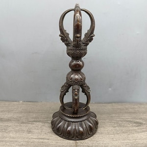 May include: A bronze-colored, ornate Vajra, a ritual object with a central sphere, decorative bands, and a three-pronged top. The base is a lotus flower design. The object is approximately 8 inches tall.