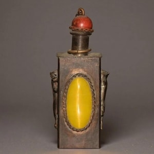 Pode incluir: Uma garrafa de rapé retangular de metal com uma incrustação central de pedra amarela ovalada rodeada por uma borda de corda. A garrafa tem figuras de animais metálicas em ambos os lados e um pescoço cilíndrico com uma tampa de pedra vermelha.