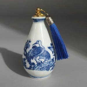 Könnte beinhalten: Eine kleine weiße Keramikflasche mit einem blau bemalten Vogel- und Blumenmuster. Die Flasche hat einen goldfarbenen Metallverschluss mit einer Öse und einer langen, leuchtend blauen Stoffquaste, die oben als dekoratives Element befestigt ist.