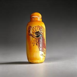 以下が含まれることがあります： 丸みを帯びた上部と別の蓋が付いた、小さな黄色のボトル。茶色と緑のアクセントが付いたカブトムシの詳細なイラストが、枝にとまっています。ボトルの高さは約10cmです。