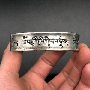 Puede incluir: Un brazalete plateado con escritura grabada. El brazalete tiene una superficie ligeramente texturizada y una inscripción oscura que envuelve la banda. El brazalete se sostiene en una mano sobre un fondo oscuro.