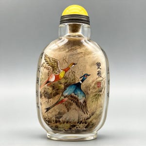 May include: A clear glass snuff bottle featuring an interior painting of two birds in flight, one with orange and yellow plumage and the other with blue and brown feathers. The bottle has a bright yellow stopper and black Chinese calligraphy characters.