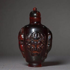 May include: A dark brown, translucent snuff bottle carved from a single piece of material. The front features a detailed relief carving of a stylised face with prominent features, while the sides display additional relief carvings and a stopper.