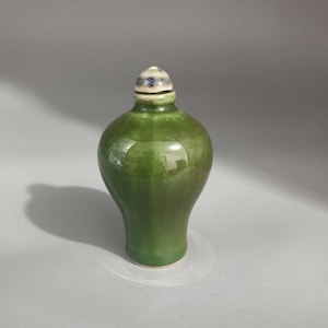 May include: A small, bulbous ceramic snuff bottle with a glossy, deep green glaze. The bottle features a narrow neck and a rounded, dome-shaped stopper that is white with blue and grey mottled accents at the top.