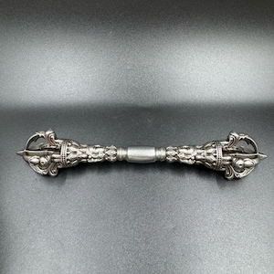 Könnte beinhalten: Ein silberfarbenes Vajra-Ritualobjekt aus Metall mit einem zentralen zylindrischen Griff, der von symmetrischen, verzierten Enden flankiert wird. Jedes Ende zeigt komplexe geschnitzte Details, einschließlich stilisierter Gesichter und gebogener Zacken.