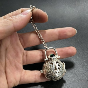 Puede incluir: Un colgante esférico de metal plateado con intrincados patrones de volutas caladas y una pequeña piedra cabujón verde en la parte superior. El colgante está unido a un eslabón de cadena de metal plateado, creando un adorno colgante.