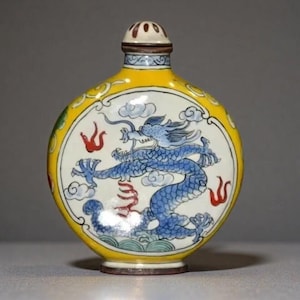 May include: A round ceramic snuff bottle featuring a yellow border and a central white panel decorated with a blue dragon, light blue clouds, and red flame motifs. The bottle has a small, matching stopper with a brown rim and a white top.
