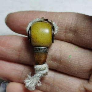 Puede incluir: Un pequeño colgante con una cuenta lisa de color amarillo mostaza en la parte superior, unida a una base cónica de color marrón oscuro mediante una banda metálica plateada con grabados decorativos. Un cordón trenzado grueso.