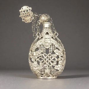 Puede incluir: Frasco de perfume plateado adornado con un intrincado diseño calado. El frasco presenta un tapón conectado por una cadena y patrones detallados de nudos, nubes y formas geométricas. El frasco tiene forma de lágrima y una parte superior decorativa.
