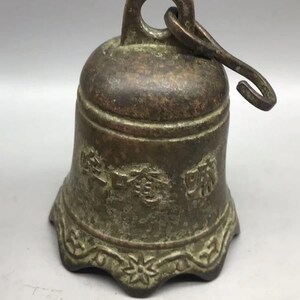 May include: A weathered bronze-toned metal bell featuring a textured surface with embossed Asian characters around the center and a decorative floral pattern along the scalloped bottom edge. A metal hook is attached to the top loop for hanging.