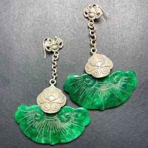 May include: A pair of drop earrings featuring silver-toned metal hooks and chain links connected to ornate silver-toned decorative caps. Each earring displays a large, fan-shaped green stone pendant with etched line details radiating outward.
