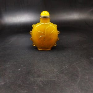 以下が含まれることがあります： 前面に鳥の彫刻が施された、小さく丸い半透明の黄色の嗅ぎタバコ入れ。ボトルの外縁には小さな丸い突起が並んでおり、ロープのような質感の金属製のキャップが付いています。