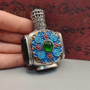 May include: A small, ornate silver bottle with a green gemstone stopper and a decorative blue and red floral design. The bottle has a detailed filigree crown and a green gemstone on the side. The bottle is approximately 7.6 cm tall.