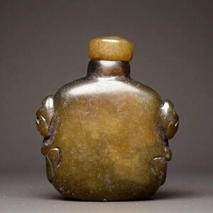 May include: A small, smooth, dark olive-green snuff bottle carved from stone. The bottle features a rounded, rectangular body with two sculpted animal-like figures on the sides and a matching cylindrical stopper at the top of the narrow neck.