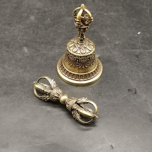 May include: A brass-colored metal bell with an ornate handle and a matching brass-colored metal vajra. The bell features intricate embossed patterns around its body and a decorative top. The vajra has a symmetrical design with curved prongs.