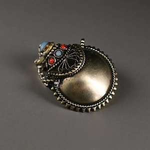 May include: A silver-toned pendant with a circular design. The pendant features a raised central area, a decorative border, and an ornate top section with red, blue, and turquoise accents. The pendant has a loop for hanging.