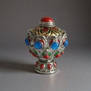 May include: An ornate silver bottle with a red top and base. The bottle is decorated with blue, red, and green accents. The design includes intricate patterns and a detailed, jewel-like appearance. A unique, handcrafted item.