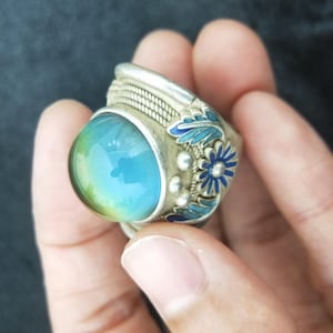 May include: A silver-toned ring featuring a large, smooth, dome-shaped cabochon stone that transitions from blue to green. The metal band is decorated with intricate silver wire patterns and small blue enamel floral and leaf motifs.
