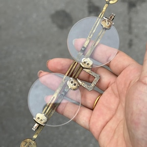 May include: Antique-style pince-nez glasses featuring round clear lenses held by a tarnished brass frame. The bridge and lens clips are decorated with small skull motifs and intricate engraved patterns, connected by a vertical metal bar structure.