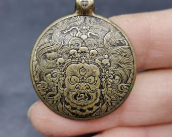 May include: A circular brass-toned metal pendant featuring an intricate embossed design of a fierce deity with a third eye, wide eyes, and an open mouth, surrounded by swirling patterns and a small bail at the top for hanging on a chain or cord.