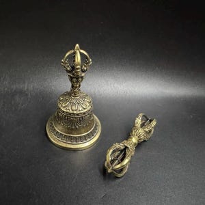 May include: A brass-coloured metal bell with an ornate handle featuring a vajra design, accompanied by a separate matching brass-coloured metal vajra tool. Both items display intricate embossed patterns and a weathered, antique-style finish.